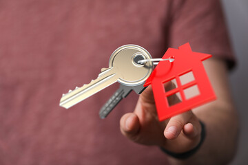 A person's hand offering a house key with a house-shaped charm, symbolizing buying, owning, or renting a property. Represents the feeling of sec