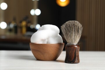Shaving brush and bowl of foam on light wooden table in barbershop, closeup