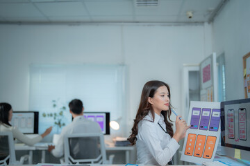 Young Asian woman UI UX designer analyzing and discussing mobile application interface prototypes with colleagues in a modern office, developing technology solutions