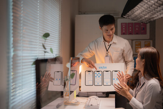 Two Asian colleagues collaborating on user experience and user interface design, presenting mobile application wireframes to optimize digital product functionality in the office