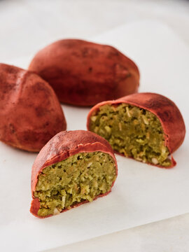 Freshly baked sweet potato pastries with a vibrant red exterior, featuring a gooey green almond filling, are arranged on a clean white surface, highlighting their texture and color