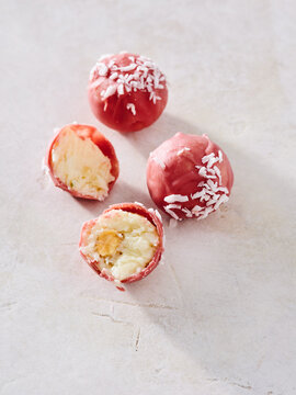 Four colorful coconut truffles are arranged on a light surface, with one truffle cut open to reveal its creamy interior. The pink coating and shredded coconut add a delightful touch