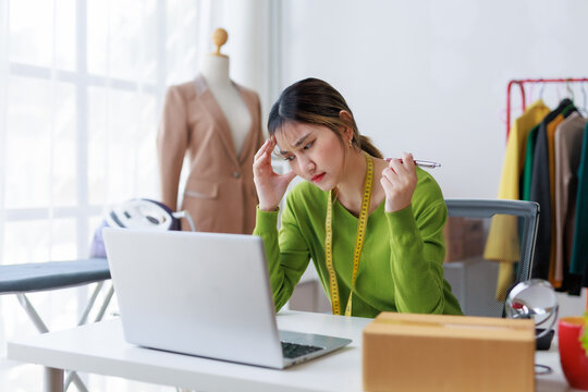 Asian female fashion designer having headache from work stress, using laptop at her studio, dealing with small business challenges