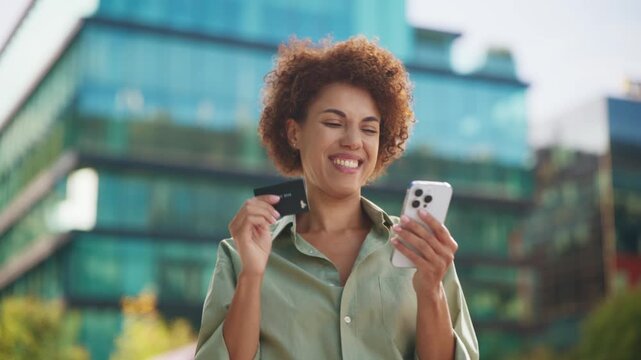 Smiling African American woman making online purchase outdoors near modern office building. Female holding credit card and smartphone entering payment information with cheerful expression outside.