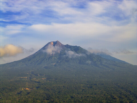 Aerial view of the majestic Merapi Mountain, cloaked in emerald forests and crowned with a smoky peak under a sky of azure and cotton clouds, Klaten, Central Java, Indonesia.