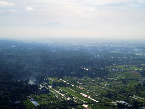 Aerial view of a sprawling landscape where verdant rice paddies meet dense forests under a hazy sky, with Mount Merapi looming in the distance, Klaten, Central Java, Indonesia.