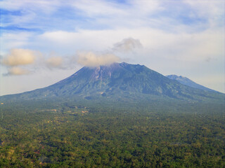 Aerial view of Merapi Mountain rising majestically above a dense forest, its peak veiled in soft clouds, a symphony of green and grey, Klaten, Central Java, Indonesia.