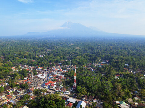 Aerial view of the red and white tower standing tall amidst a vibrant tapestry of green trees, with Merapi Mountain looming in the distance, Klaten, Central Java, Indonesia.