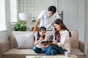 Asian parents and daughter sitting on sofa, connecting with a digital tablet for online learning, bonding, and remote education