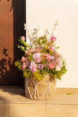 A vibrant flower arrangement featuring pink roses, hydrangeas, and delicate blossoms sits inside a woven basket on a step. Warm sunlight casts beautiful shadows, enhancing the scene.