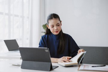 Asian woman focusing on her book, researching and studying while working from a modern workspace with digital devices