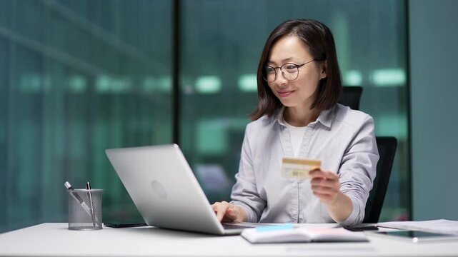 Happy asian businesswoman doing online shopping typing credit card data on laptop computer while sitting in business office. Smiling female employee makes purchase, enjoy e-commerce e-shopping
