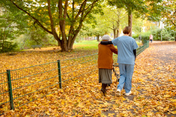 Obraz premium Elderly woman taking a walk through the park in autumn with help of walker and caretaker aid