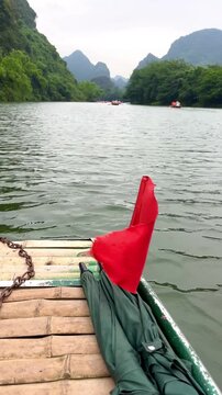 Vertical 4K video of a quiet boat trip with Vietnamese flag on the Trang An river in Ninh Binh surrounded by beautiful mountains and lush greenery outdoor at daytime in Vietnam while traveling in Asia