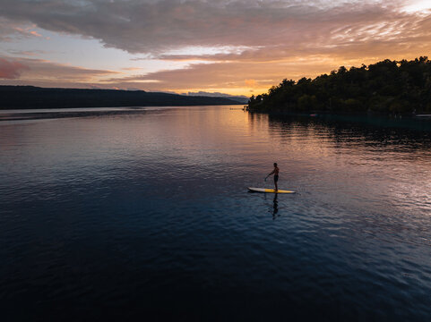 Aerial view of a lone paddleboarder glides across the serene waters, reflecting the vibrant sunset hues near the verdant shores of Moso Island, Moso island, Shefa Province, Vanuatu.