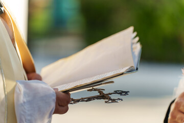In a serene outdoor location, a person dressed in formal attire holds an open book and a cross,...