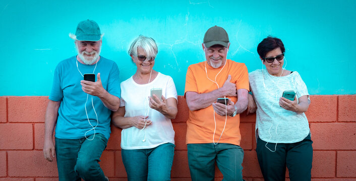 Group of four happy senior friends standing against a colorful wall using smartphones and listening to music on headphones. A concept of active aging, friendship, digital connection among seniors