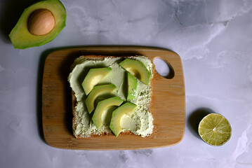 square table of bread with spread avocado butter with avocado slices on wooden board on dark background