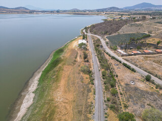 Aerial view showcasing a road and railway alongside Valencia Dam in Jalisco, Mexico. Parched landscape, agave plantations, and distant mountains under a bright, clear sky
