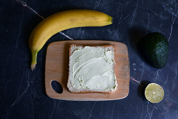square table of bread with spread avocado butter with avocado slices on wooden board on dark background