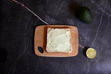square table of bread with spread avocado butter on wooden board on dark background
