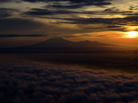 Aerial view of a lawu mountain range piercing through a thick blanket of clouds as the sun rises, casting a warm golden glow across the horizon, Karanganyar Regency, Central Java, Indonesia.