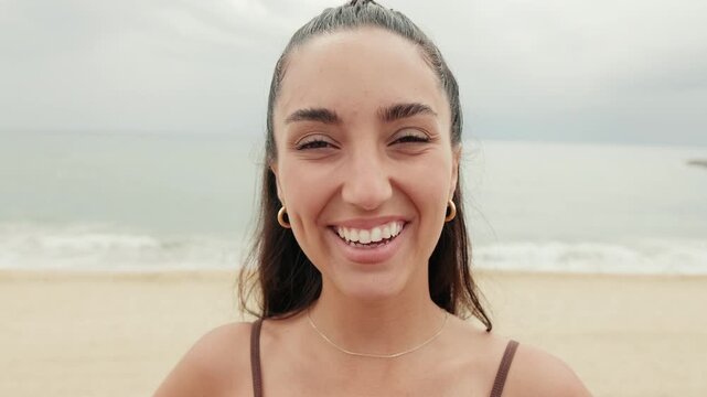 Young woman smiles at the beach on an overcast day. She has dimples and is wearing a brown bathing suit top.