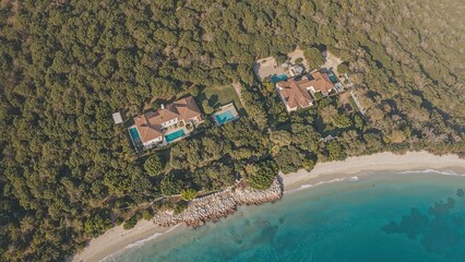 Secluded luxury villas with swimming pools nestled within a fragrant pine forest overlooking pristine beachfront, captured from above showcasing the charming real estate of a Mediterranean island.