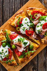 Pinsa with cured ham, burrata cheese, tomatoes, green olives and arugula on wooden table	