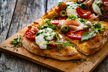 Pinsa with cured ham, burrata cheese, tomatoes, green olives and arugula on wooden table	
