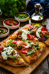 Pinsa with cured ham, burrata cheese, tomatoes, green olives and arugula on wooden table	