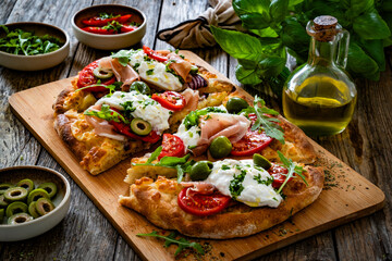 Pinsa with cured ham, burrata cheese, tomatoes, green olives and arugula on wooden table	