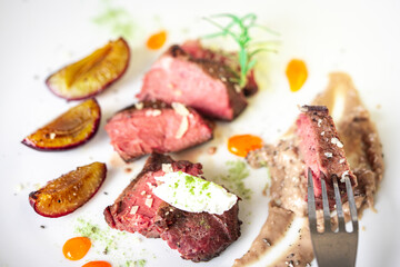 Medium-rare steak, sliced, with dogwood sauce on a white plate with roasted plums and rosemary cream cheese dumplings. Top view.
