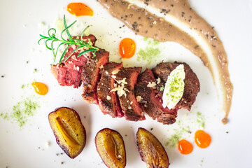 Medium-rare steak, sliced, with dogwood sauce on a white plate with roasted plums and rosemary cream cheese dumplings. Top view.