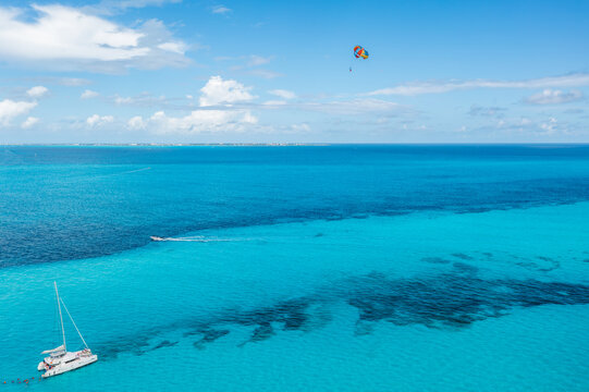 Aerial view of the turquoise sea contrasting with the white catamaran and distant city, a parasail adding a splash of color against the blue sky, Cancun, Mexico.