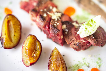 Medium-rare steak, sliced, with dogwood sauce on a white plate with roasted plums and rosemary cream cheese dumplings. Top view.