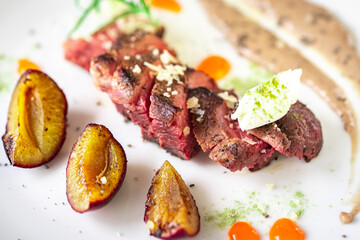 Medium-rare steak, sliced, with dogwood sauce on a white plate with roasted plums and rosemary cream cheese dumplings. Top view.