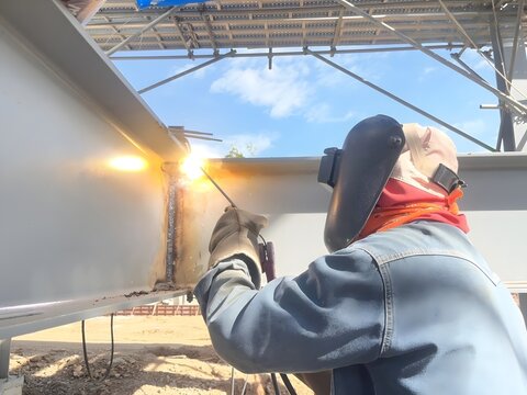 Industrial welder working outdoors on steel beam construction, professional welding sparks scene, metalwork, craftsmanship, safety equipment, heavy industry, engineering, and skilled labor concept