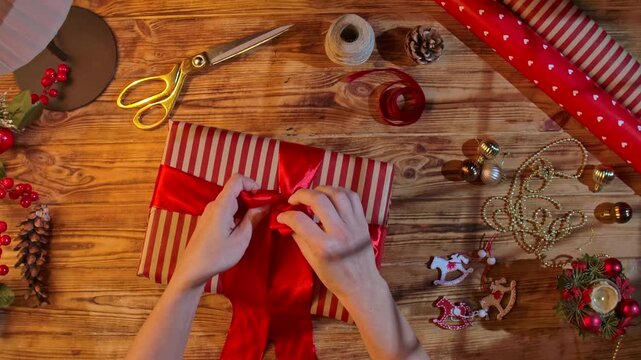 Top view of a woman hands tying a red satin ribbon on a striped gift box among scissors, twine, and decorations on a rustic wooden table for Christmas crafts.