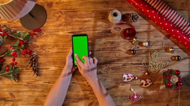 Top view of hands swiping a smartphone with a green screen on a rustic wooden table surrounded by Christmas decorations and wrapping materials, ideal for festive shopping or design layouts.