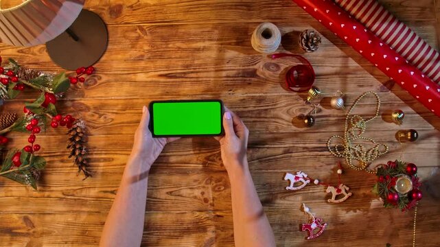 Top view of hands holding a smartphone with a green screen on a rustic wooden table surrounded by Christmas decorations and wrapping materials, ideal for festive shopping or design layouts.