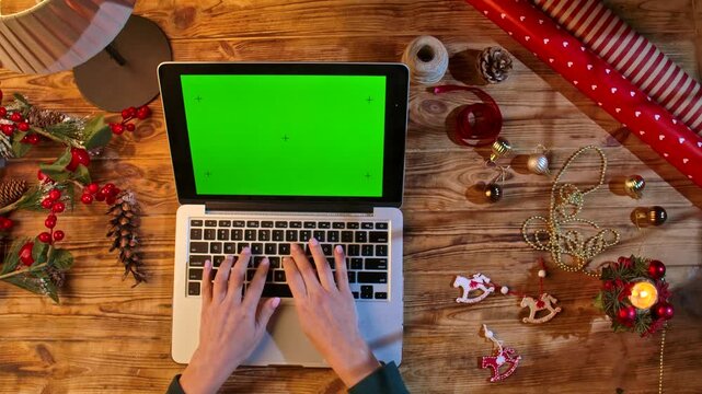 Top view of hands typing on a laptop with a chromatic green screen on a wooden table surrounded by packaging materials and decorations. The perfect layout for holiday shopping or design.