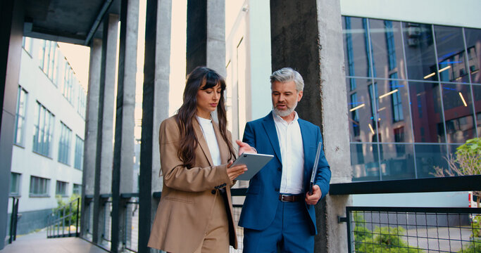 Confident business professionals in formal suits walking and talking outside a modern office building female executive using a digital tablet, male colleague holding a laptop