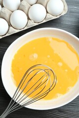 Beaten eggs in bowl and whisk on dark wooden table, flat lay