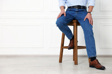Man in stylish jeans on stool near white wall, closeup. Space for text