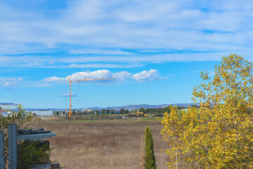 Yellow tower crane working on open construction site in rural field with distant buildings. Concept of land development, suburban expansion, construction progress, urban sprawl