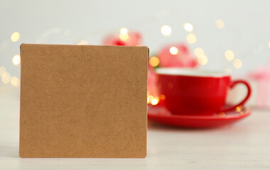 Blank card and cup of coffee on white wooden table against blurred background, bokeh effect. Mockup for design