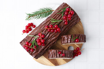 Delicious Yule log (traditional Christmas cake) with redcurrant and fir tree branch on white tiled table, top view