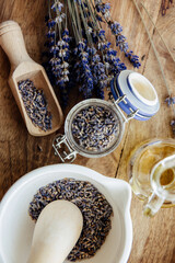 Dried lavender flower buds inside clear glass jar, with mortar and pestle filled with flower and bouquet next to it. Brown wood background.