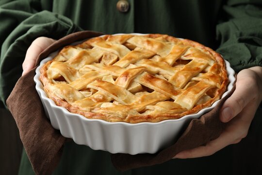 Woman with baking dish of delicious homemade apple pie against dark background, closeup - Powered by Adobe